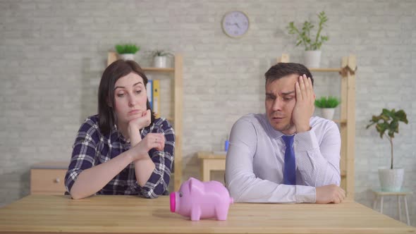 Married Couple Sits at Home at the Table and Looks Puzzled at the Piggy Bank