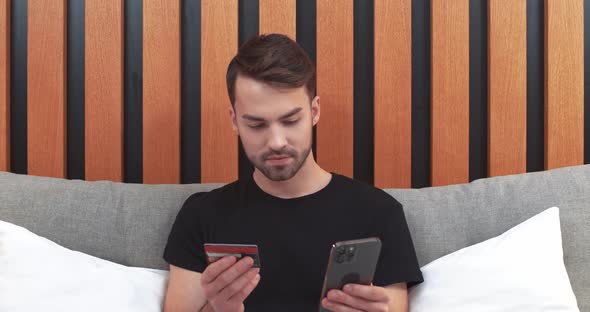 Young Man Sitting on the Bed and Pays for Purchases Online Using a Smartphone Enters Credit Card