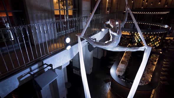 Female aerialist doing a front split turn on white silks above a bar.