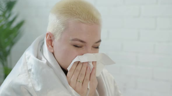 Upset Woman with Short White Hair Wrapped in White Blanket at Home Blows Her Runny Nose Into