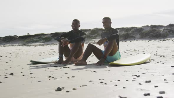 Happy african american teenage twin brothers sitting by surfboards on a beach talking