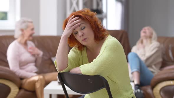 Displeased Redhead Woman Sitting on Chair Shaking Head Talking As Blurred Cheerful Friends Laughing