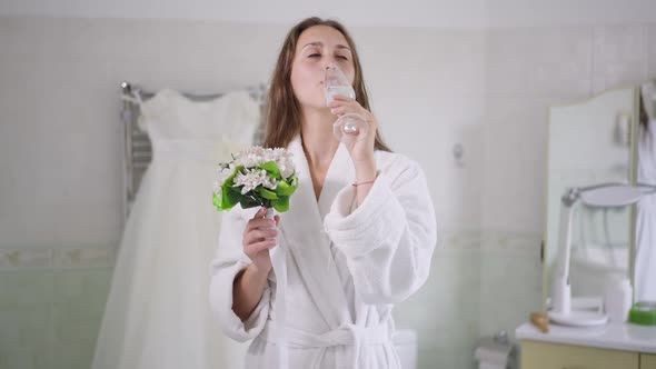 Front View of Smiling Charming Young Caucasian Bride Drinking Champagne Smelling Bouquet of Flowers