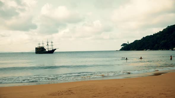 A medieval ship is getting closer to the beach to dock on the port on a cloudy day with people on th