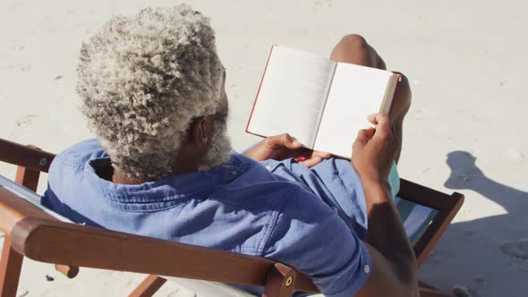 Senior african american man reading and lying on sunbed on sunny beach