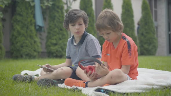 Brunette Boy with Tablet Talking with Cute Little Kid Holding Headphones. Wide Shot Portrait of