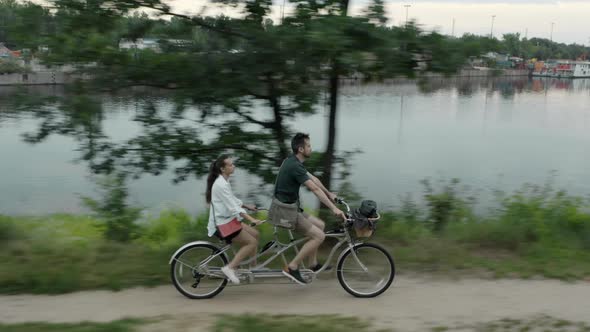 A young couple rides a tandem two-seater bike along the riverbank