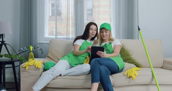 Two Women-Cleaners in Uniforms Sitting on Couch in Living-Room After Cleaning up Client's Flat