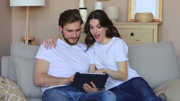 Happy young couple sitting, relaxing on sofa in living room watching media content online