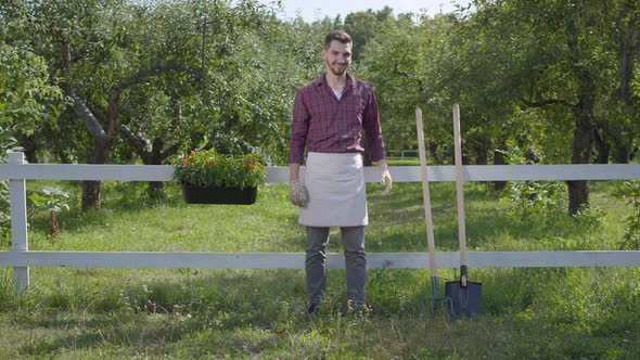 Professional Young Farmer in Garden Gloves Standing in the Green Summer Garden Near the Fence