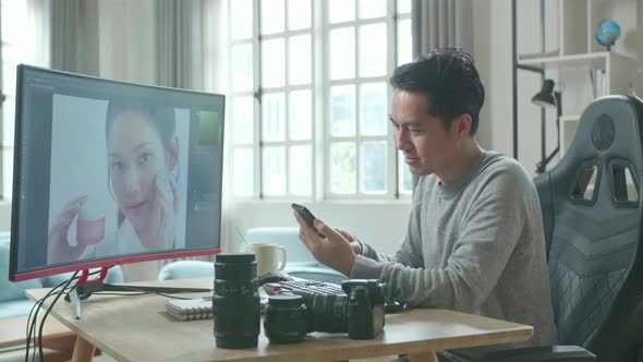 Asian Cameraman Using Mobile Phone While Using Desktop Computer For Working At Home