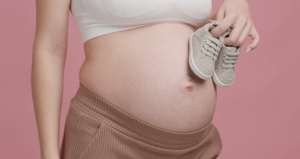 Close Up Shot of Pregnant Woman Holding Tiny Boots Near Her Big Belly Pink Studio Background