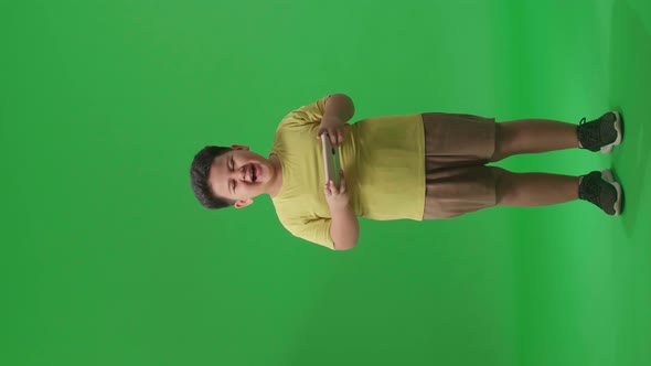 Full Body Of Laughing Asian Little Boy Looking At The Phone On Green Screen In The Studio