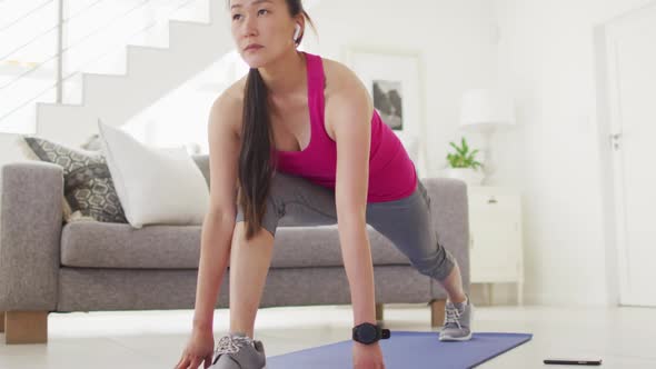 Asian woman on mat wearing earphones, exercising with smartphone at home