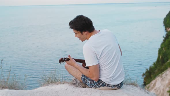 Young Smiling Man Playing Ukulele and Singing  Sitting on the Mountain