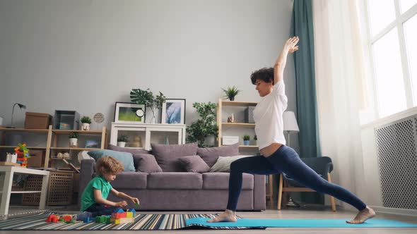 Modern Housewife Doing Yoga While Her Little Child Playing with Blocks on Carpet