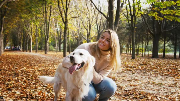 Young Female in Casual Clothes is Squatting on Alley of Autumn Park Smiling and Stroking Her Dog