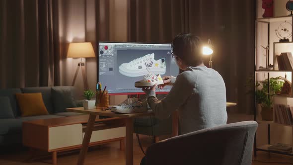 Asian Male Footwear Designer Looking At Sneaker In Hands And Comparing It To Desktop At Home