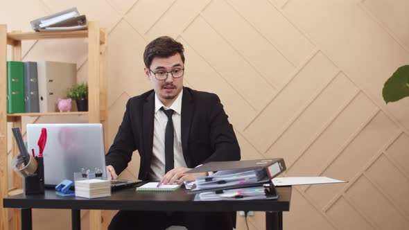 Man in Suit and Glasses is Sitting Table with Laptop and Looking Tired Eyes at Folders with Papers