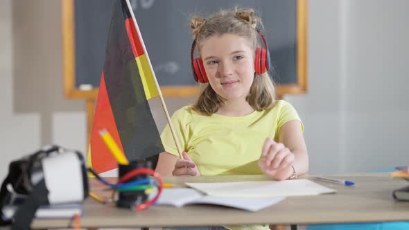 Portrait of Charming Smiling German Schoolgirl in Headphones Sitting at Desk in Classroom Holding