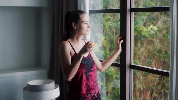 Joyful Woman in Elegant Pajama Drinks Juice Near Window