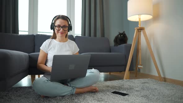 Casually Dressed Woman with Headphones is Sitting on Carpet with Laptop and Works Remotely in Cozy