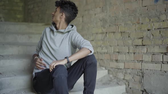 Portrait of Troubled Young Man Sitting on Stairs in Abandoned Building and Looking Around. Stressed