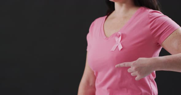 Mid section of a woman pointing to the pink ribbon on her chest against black background