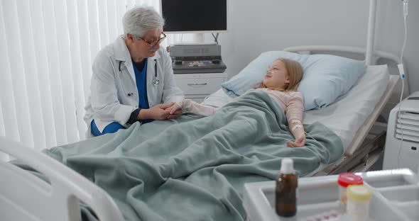 Senior Male Doctor Holding Hand and Talking To Sick Girl Patient Resting in Hospital Bed