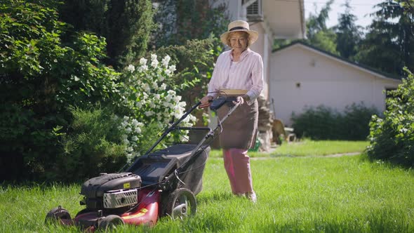 Wide Shot Portrait of Positive Senior Woman Gesturing Thumb Up Smiling Standing with Lawn Mower