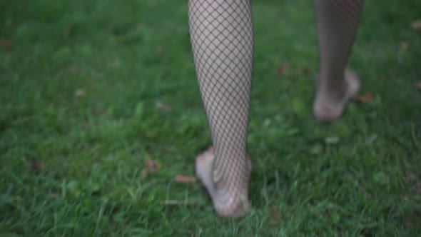 Tracking Shot Barefoot Slim Young Drunken Woman in Fishnet Stockings Walking on Green Grass Passing