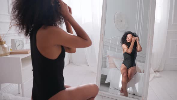 Black Woman Strokes Her Hair Sitting on a Bed and Looking in Mirror Hair Care