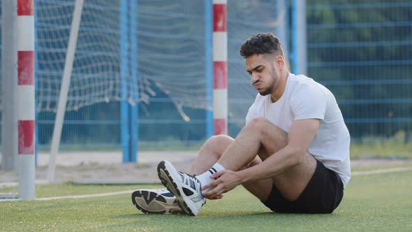 Painsuffering Middle Eastern Footballer Sitting on Grass of Soccer Field Against Goal Holding on