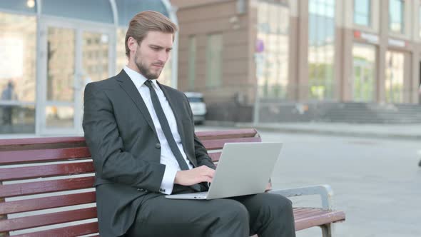 Young Businessman with Wrist Pain Using Laptop While Sitting on Bench