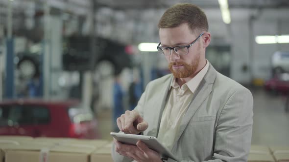 Portrait of Redhead Caucasian Man in Eyeglasses Making Deal Online Using His Tablet, Looking at