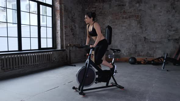 Focused young Indian woman cycling in gym. Side view of slim brunette sportswoman training