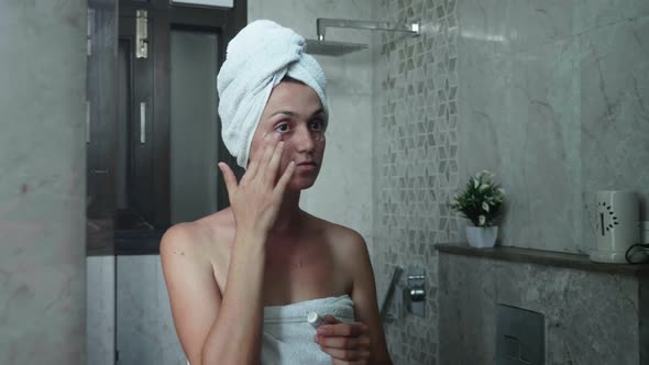 Young Woman is Applying Cream on the Face Eye Area While Looking at Mirror