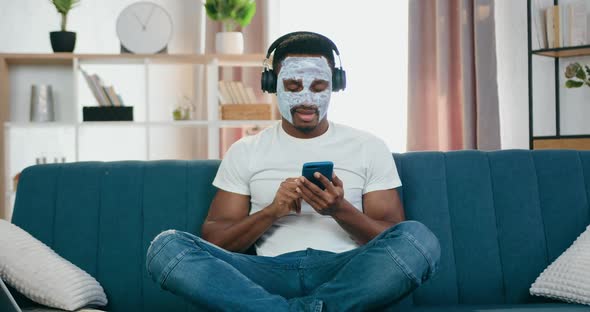 African American with Facial Mask Sitting in Lotus Pose on the Couch and Enjoying Favourite Music