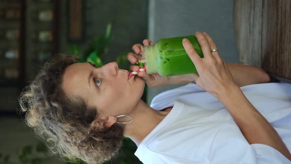 Vertical Shot of Young Curly Woman Drinking Fresh Green Juice Sitting in Trendy Cafe Enjoying