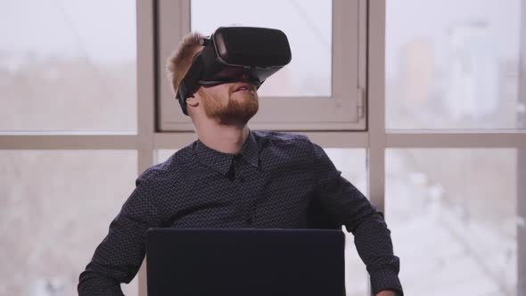 Man Executive Using Virtual Reality Headset Sitting at Desk with Laptop in Office.