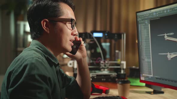 Asian Engineer Talking On Mobile Phone While Work On Computer And 3D Printer, Screen Shows 3d Drone