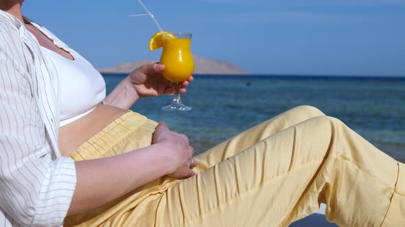 Pregnant Woman Drinking Orange Juice While Relaxing On The Beach By The Sea