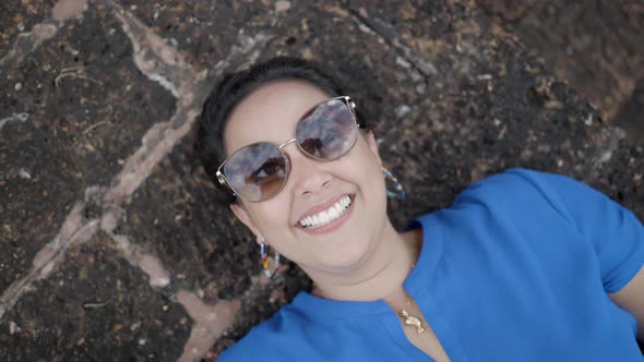 Female Tourist In Blue Shirt And Sunglasses Smiling While Lying Down On Old Concrete Bench At