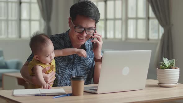 Male Holding His Son And Talking Over Phone During Work With Laptop Computer At Home