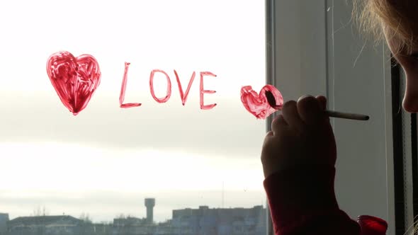 Little girl holds paintbrush in hand draws red heart on window Valentine's day love home family art