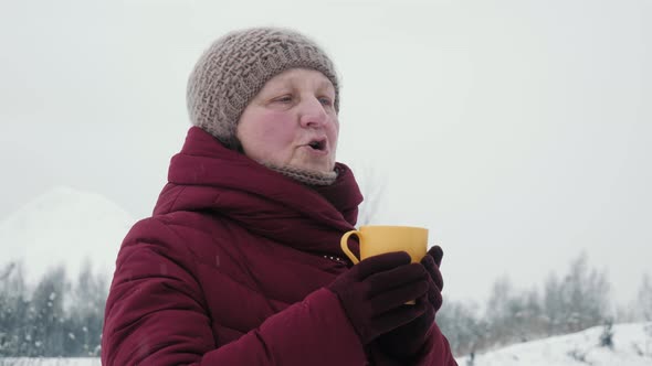 Elderly woman in warm hat is drinking hot tea or coffee from a cup in a snowy forest