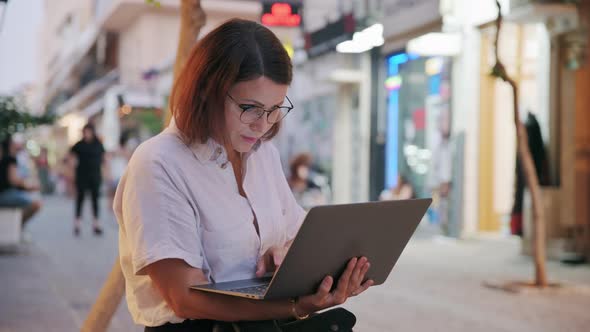 Middleaged Woman Sitting with a Laptop on a Bench in the Evening City
