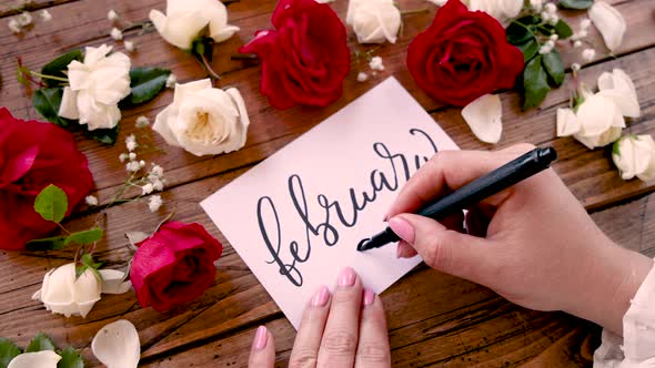 Writing FEBRUARY and drawing heart on a card near flowers close up