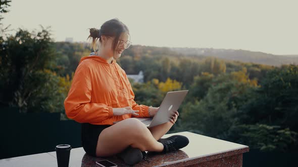 The Student with Glasses and Orange Blouse Sitting in the Park Talks By Video Call with Her Parents