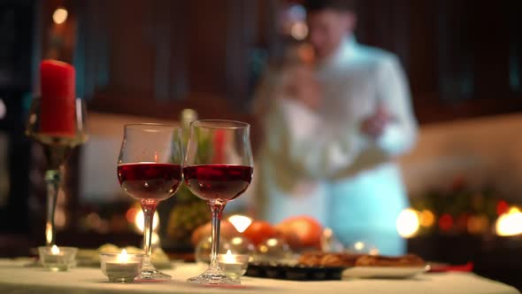 Closeup Red Wine Glasses on Table with Blurred Loving Adult Couple Dancing at Background in Kitchen